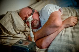 homme endormi les bras croisés avec masque respiratoire branché à une machine de traitement des troubles du sommeil
